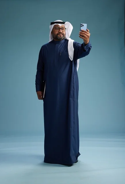 رجل سعودي برتدي ثوب ازرق وغتره بيضاء يبتسم ويلتقط صورة سيلفي باستخدام الهاتف الذكي, اجراء مكاملة فيديو عبر الجوال, كامل الجسم, خلفيه زرقاء