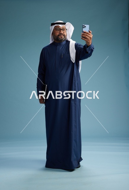 رجل سعودي برتدي ثوب ازرق وغتره بيضاء يبتسم ويلتقط صورة سيلفي باستخدام الهاتف الذكي, اجراء مكاملة فيديو عبر الجوال, كامل الجسم, خلفيه زرقاء