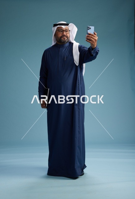 رجل سعودي برتدي ثوب ازرق وغتره بيضاء يبتسم ويلتقط صورة سيلفي باستخدام الهاتف الذكي, اجراء مكاملة فيديو عبر الجوال, كامل الجسم, خلفيه زرقاء