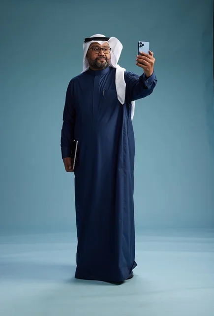 A Saudi man wearing a blue thobe and a white ghutrah is smiling and taking a selfie using a smartphone, making a video call via mobile, full body, with a blue background.