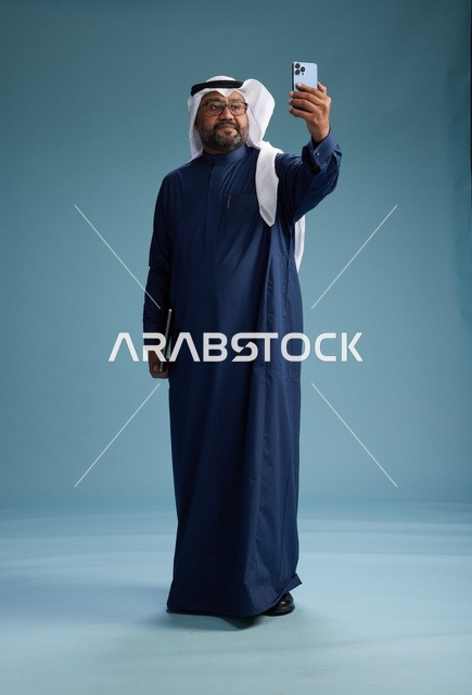 رجل سعودي برتدي ثوب ازرق وغتره بيضاء يبتسم ويلتقط صورة سيلفي باستخدام الهاتف الذكي, اجراء مكاملة فيديو عبر الجوال, كامل الجسم, خلفيه زرقاء