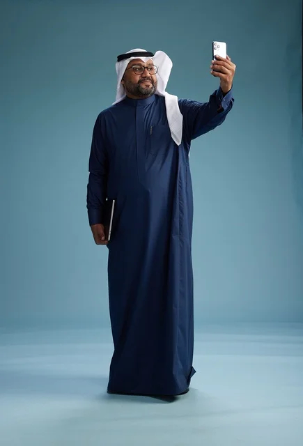 A Saudi man wearing a blue thobe and a white ghutrah is smiling and taking a selfie using a smartphone, making a video call via mobile, full body, with a blue background.
