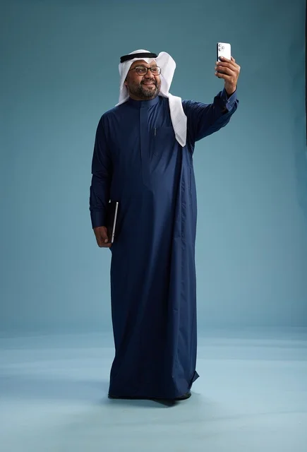 A Saudi man wearing a blue thobe and a white ghutrah is smiling and taking a selfie using a smartphone, making a video call via mobile, full body, with a blue background.