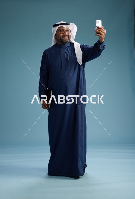 A Saudi man wearing a blue thobe and a white ghutrah is smiling and taking a selfie using a smartphone, making a video call via mobile, full body, with a blue background.
