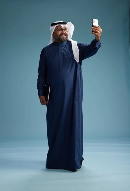 A Saudi man wearing a blue thobe and a white ghutrah is smiling and taking a selfie using a smartphone, making a video call via mobile, full body, with a blue background.