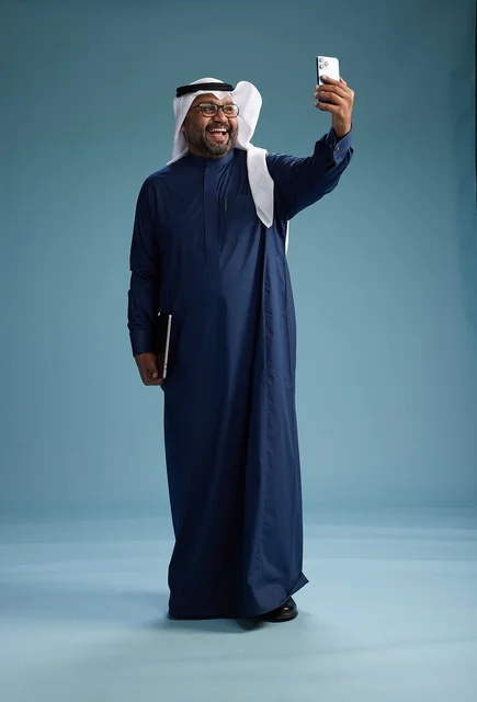 A Saudi man wearing a blue thobe and a white ghutrah is smiling and taking a selfie using a smartphone, making a video call via mobile, full body, with a blue background.