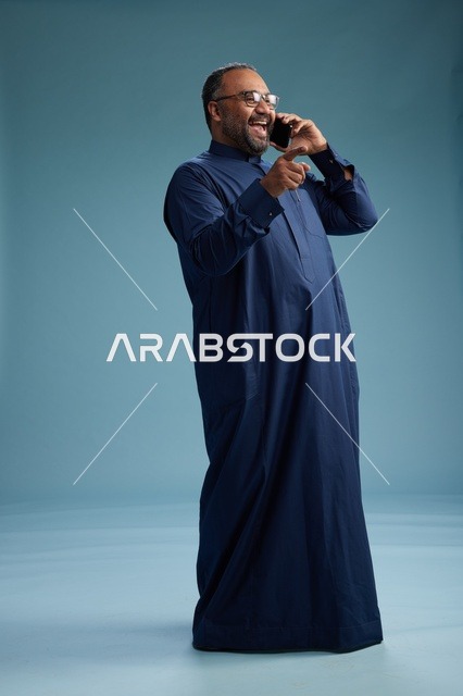 بورتريه لرجل سعودي يرتدي ثوب كحلي في مكالمة هاتفية, دردشه ومحادثات عبر الجوال, الوقوف بالطول الكامل, خلفيه زرقاء