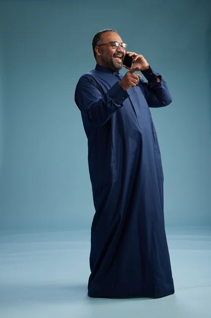 A portrait of a Saudi man wearing a navy blue thobe on a phone call, chatting and conversing via mobile, standing full-length, with a blue background.