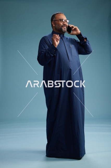 بورتريه لرجل سعودي يرتدي ثوب كحلي في مكالمة هاتفية, دردشه ومحادثات عبر الجوال, الوقوف بالطول الكامل, خلفيه زرقاء