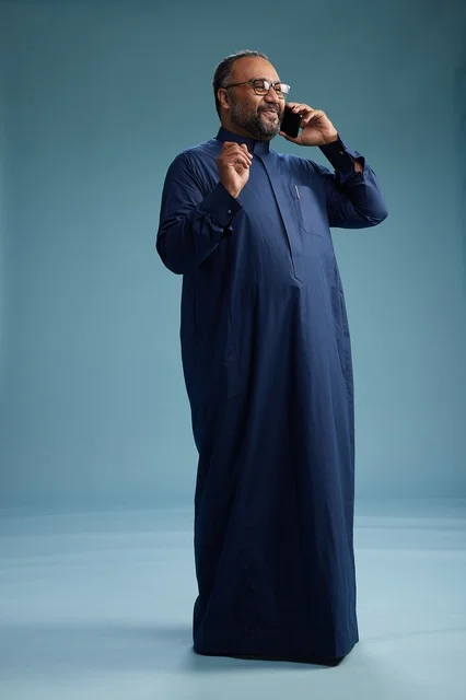 A portrait of a Saudi man wearing a navy blue thobe on a phone call, chatting and conversing via mobile, standing full-length, with a blue background.