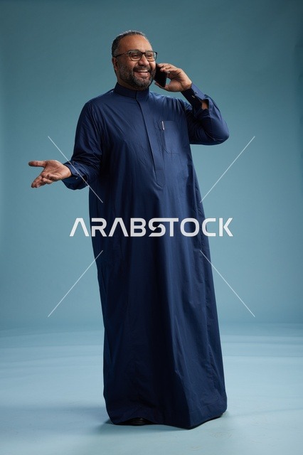 A portrait of a Saudi man wearing a navy blue thobe on a phone call, chatting and conversing via mobile, standing full-length, with a blue background.