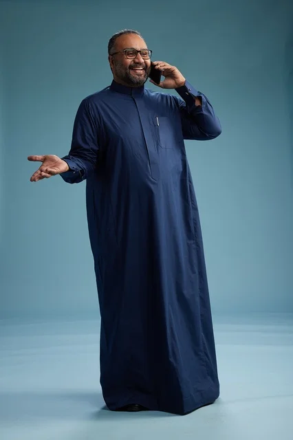 A portrait of a Saudi man wearing a navy blue thobe on a phone call, chatting and conversing via mobile, standing full-length, with a blue background.