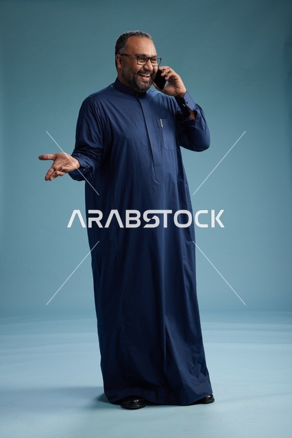 بورتريه لرجل سعودي يرتدي ثوب كحلي في مكالمة هاتفية, دردشه ومحادثات عبر الجوال, الوقوف بالطول الكامل, خلفيه زرقاء