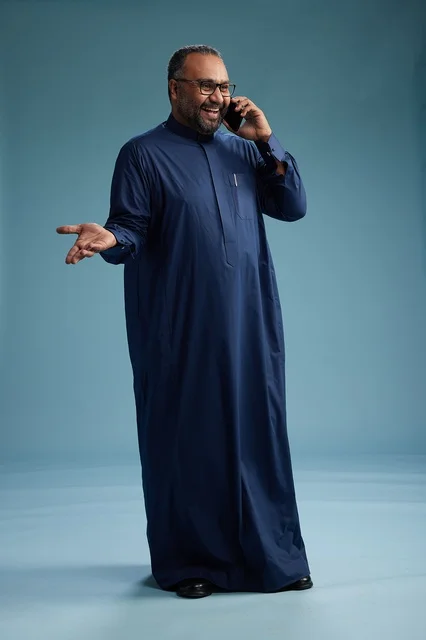 A portrait of a Saudi man wearing a navy blue thobe on a phone call, chatting and conversing via mobile, standing full-length, with a blue background.