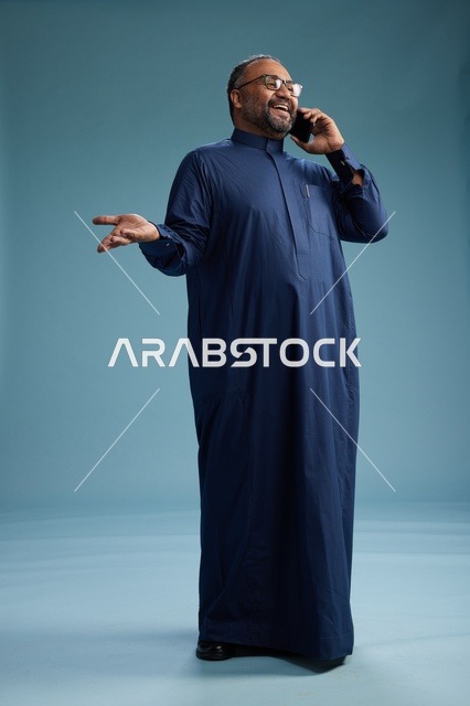 بورتريه لرجل سعودي يرتدي ثوب كحلي في مكالمة هاتفية, دردشه ومحادثات عبر الجوال, الوقوف بالطول الكامل, خلفيه زرقاء