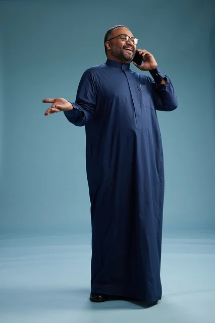 A portrait of a Saudi man wearing a navy blue thobe on a phone call, chatting and conversing via mobile, standing full-length, with a blue background.