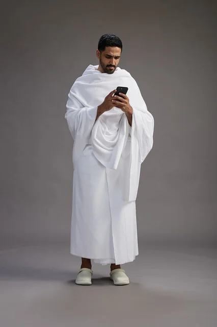 A portrait of an Arab Saudi Gulf man wearing Ihram clothing using a mobile phone, concept of Hajj and Umrah, full body, gray background.
