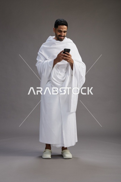 بورتريه لرجل عربي سعودي خليجي يرتدي ملابس الإحرام يستخدم الهاتف المحمول, مفهوم الحج والعمره , كامل الجسم, خلفيه رماديه