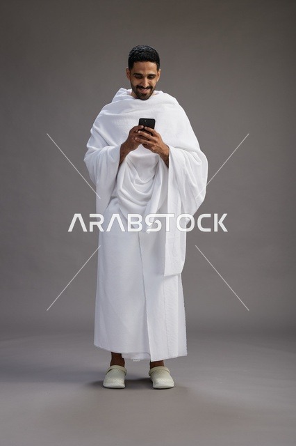 A portrait of an Arab Saudi Gulf man wearing Ihram clothing using a mobile phone, concept of Hajj and Umrah, full body, gray background.