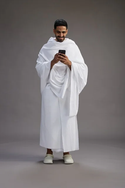 A portrait of an Arab Saudi Gulf man wearing Ihram clothing using a mobile phone, concept of Hajj and Umrah, full body, gray background.