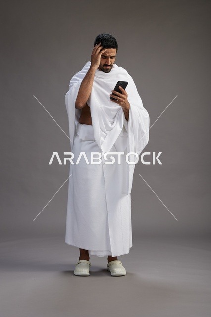 بورتريه لرجل عربي سعودي خليجي يرتدي ملابس الإحرام يستخدم الهاتف المحمول, مفهوم الحج والعمره , كامل الجسم, خلفيه رماديه