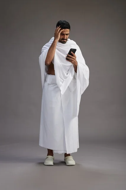 A portrait of an Arab Saudi Gulf man wearing Ihram clothing using a mobile phone, concept of Hajj and Umrah, full body, gray background.