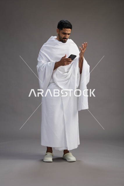 A portrait of an Arab Saudi Gulf man wearing Ihram clothing using a mobile phone, concept of Hajj and Umrah, full body, gray background.