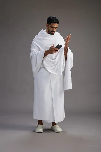 A portrait of an Arab Saudi Gulf man wearing Ihram clothing using a mobile phone, concept of Hajj and Umrah, full body, gray background.