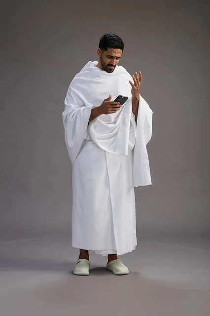 بورتريه لرجل عربي سعودي خليجي يرتدي ملابس الإحرام يستخدم الهاتف المحمول, مفهوم الحج والعمره , كامل الجسم, خلفيه رماديه
