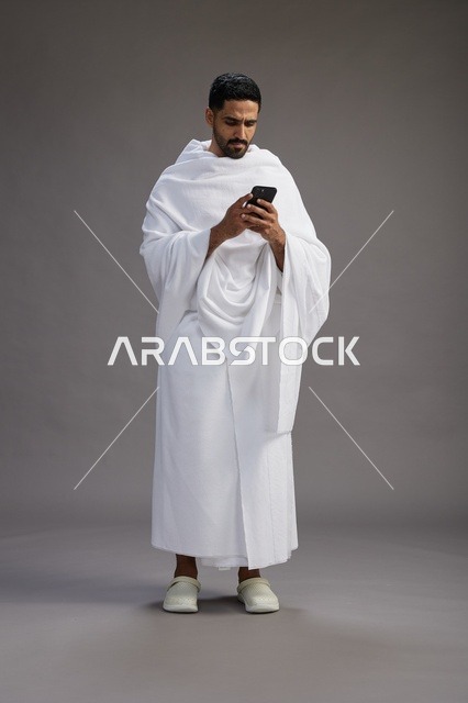 بورتريه لرجل عربي سعودي خليجي يرتدي ملابس الإحرام يستخدم الهاتف المحمول, مفهوم الحج والعمره , كامل الجسم, خلفيه رماديه