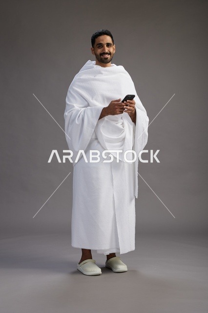 A portrait of an Arab Saudi Gulf man wearing Ihram clothing using a mobile phone, concept of Hajj and Umrah, full body, gray background.