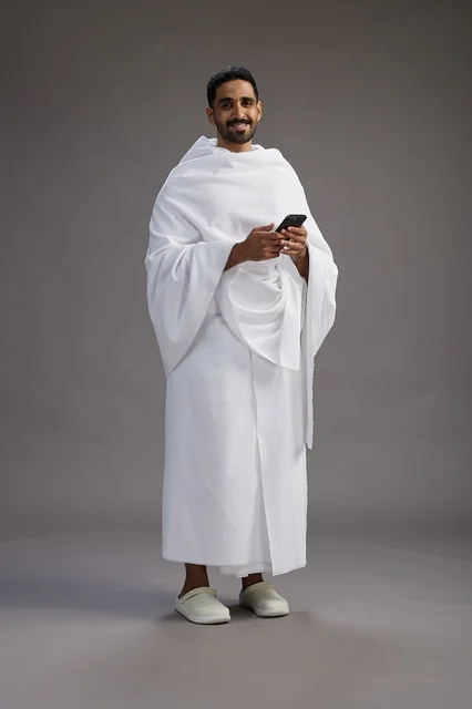 بورتريه لرجل عربي سعودي خليجي يرتدي ملابس الإحرام يستخدم الهاتف المحمول, مفهوم الحج والعمره , كامل الجسم, خلفيه رماديه