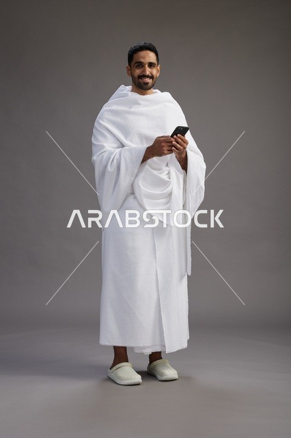 بورتريه لرجل عربي سعودي خليجي يرتدي ملابس الإحرام يستخدم الهاتف المحمول, مفهوم الحج والعمره , كامل الجسم, خلفيه رماديه