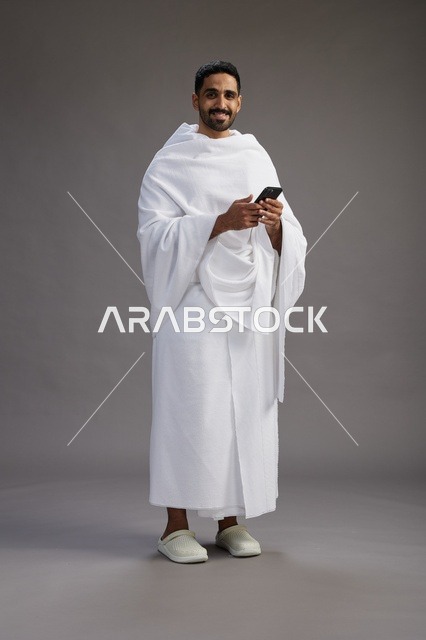 بورتريه لرجل عربي سعودي خليجي يرتدي ملابس الإحرام يستخدم الهاتف المحمول, مفهوم الحج والعمره , كامل الجسم, خلفيه رماديه