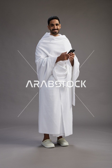 بورتريه لرجل عربي سعودي خليجي يرتدي ملابس الإحرام يستخدم الهاتف المحمول, مفهوم الحج والعمره , كامل الجسم, خلفيه رماديه