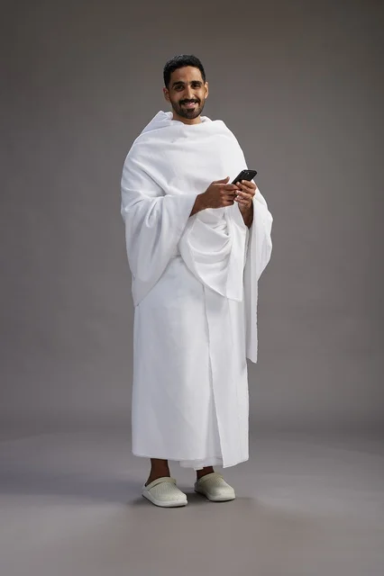A portrait of an Arab Saudi Gulf man wearing Ihram clothing using a mobile phone, concept of Hajj and Umrah, full body, gray background.