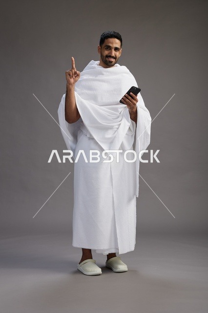 بورتريه لرجل عربي سعودي خليجي يرتدي ملابس الإحرام يستخدم الهاتف المحمول, مفهوم الحج والعمره , كامل الجسم, خلفيه رماديه