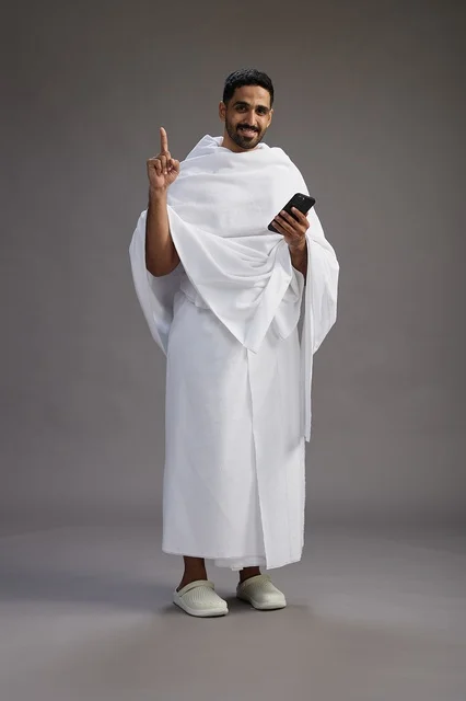 بورتريه لرجل عربي سعودي خليجي يرتدي ملابس الإحرام يستخدم الهاتف المحمول, مفهوم الحج والعمره , كامل الجسم, خلفيه رماديه