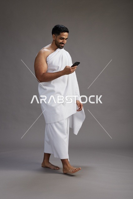 بورتريه لرجل عربي سعودي خليجي يرتدي ملابس الإحرام يستخدم الهاتف المحمول, مفهوم الحج والعمره , كامل الجسم, خلفيه رماديه