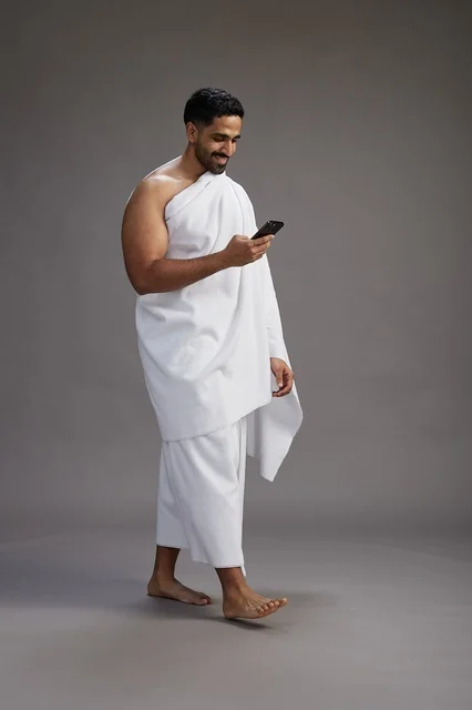 A portrait of an Arab Saudi Gulf man wearing Ihram clothing using a mobile phone, concept of Hajj and Umrah, full body, gray background.
