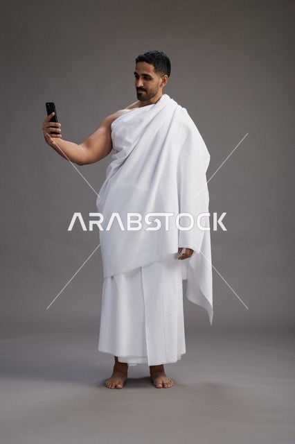 بورتريه لرجل عربي سعودي خليجي يرتدي ملابس الإحرام يستخدم الهاتف المحمول, مفهوم الحج والعمره , كامل الجسم, خلفيه رماديه