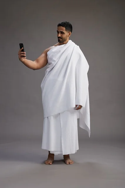 بورتريه لرجل عربي سعودي خليجي يرتدي ملابس الإحرام يستخدم الهاتف المحمول, مفهوم الحج والعمره , كامل الجسم, خلفيه رماديه