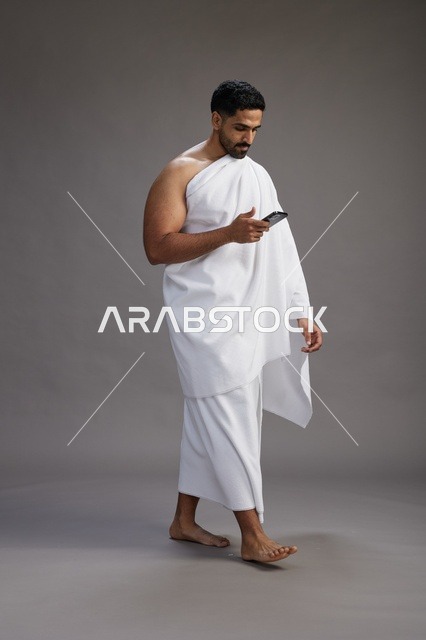 بورتريه لرجل عربي سعودي خليجي يرتدي ملابس الإحرام يستخدم الهاتف المحمول, مفهوم الحج والعمره , كامل الجسم, خلفيه رماديه