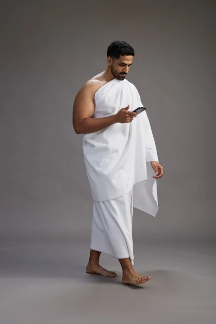 A portrait of an Arab Saudi Gulf man wearing Ihram clothing using a mobile phone, concept of Hajj and Umrah, full body, gray background.