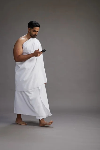 A portrait of an Arab Saudi Gulf man wearing Ihram clothing using a mobile phone, concept of Hajj and Umrah, full body, gray background.