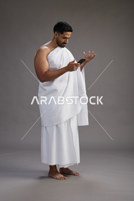 بورتريه لرجل عربي سعودي خليجي يرتدي ملابس الإحرام يستخدم الهاتف المحمول, مفهوم الحج والعمره , كامل الجسم, خلفيه رماديه