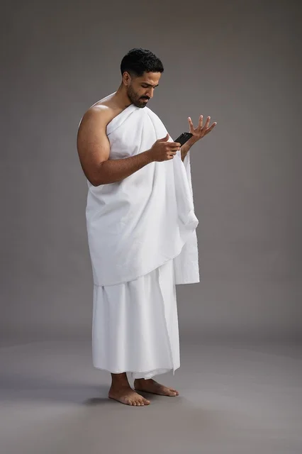 A portrait of an Arab Saudi Gulf man wearing Ihram clothing using a mobile phone, concept of Hajj and Umrah, full body, gray background.
