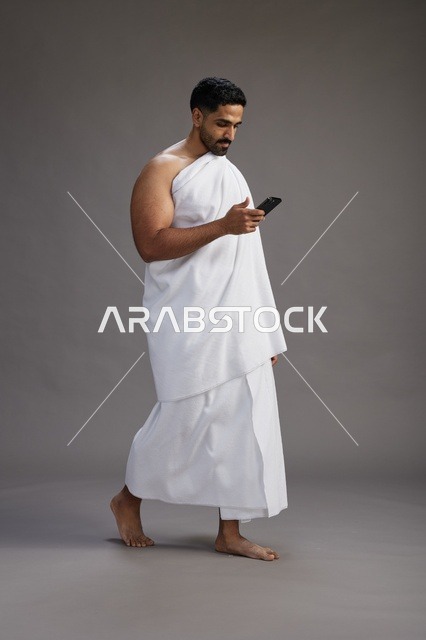 بورتريه لرجل عربي سعودي خليجي يرتدي ملابس الإحرام يستخدم الهاتف المحمول, مفهوم الحج والعمره , كامل الجسم, خلفيه رماديه