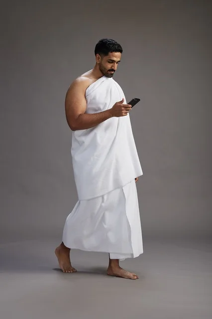 A portrait of an Arab Saudi Gulf man wearing Ihram clothing using a mobile phone, concept of Hajj and Umrah, full body, gray background.