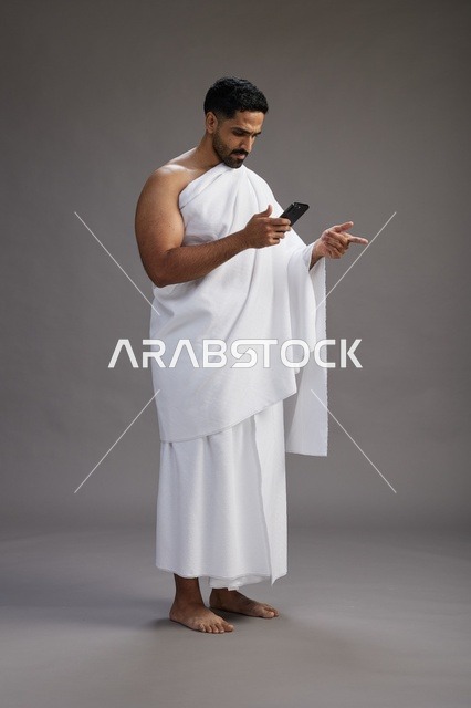 بورتريه لرجل عربي سعودي خليجي يرتدي ملابس الإحرام يستخدم الهاتف المحمول, مفهوم الحج والعمره , كامل الجسم, خلفيه رماديه